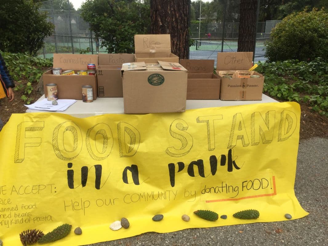 A handmade yellow Food Stand in a Park banner in front of collection boxes labeled for rice, pasta, beans, and canned food