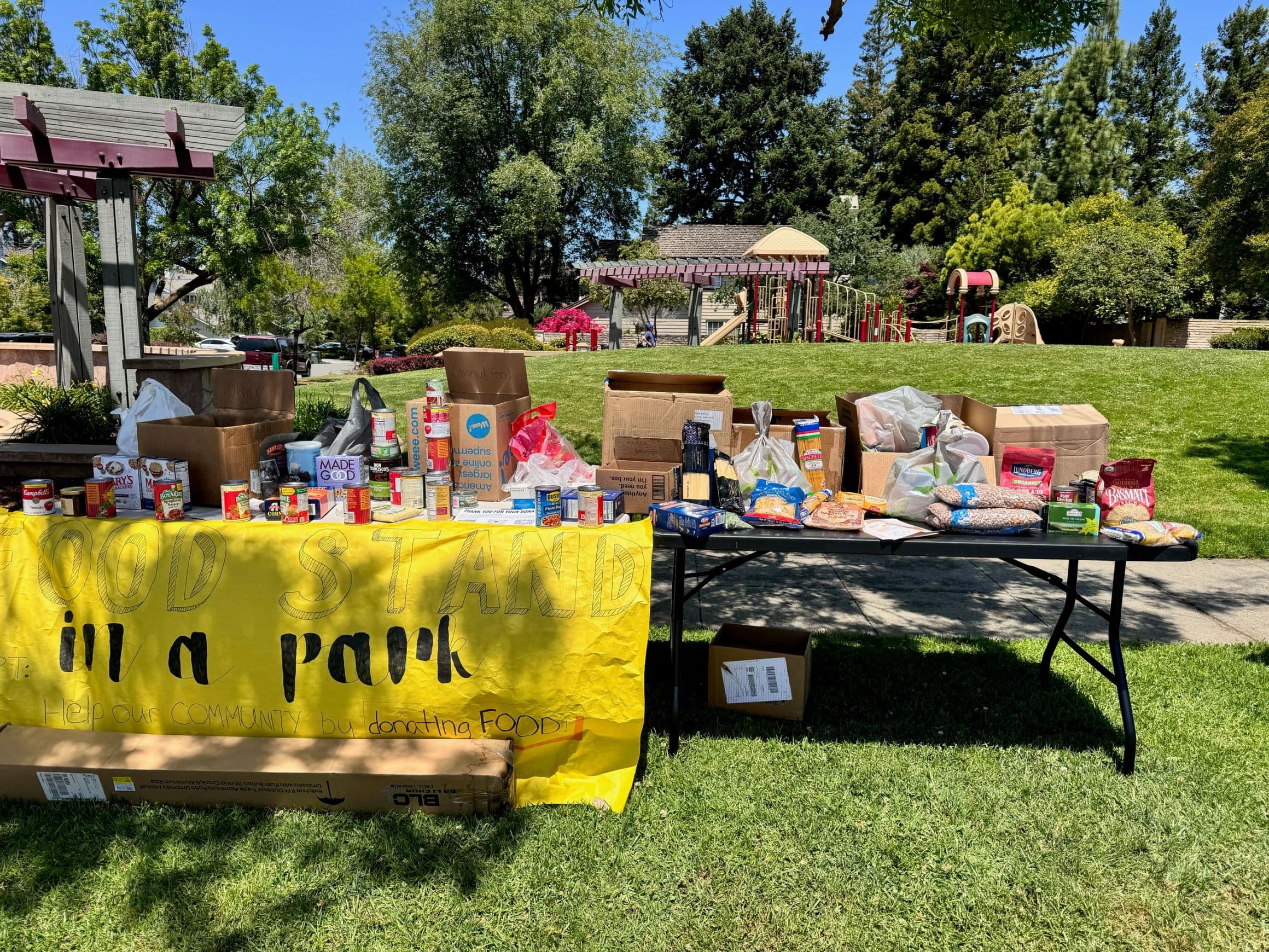Food collection tables at a park, with donated groceries and a handmade Food Stand in a Park sign